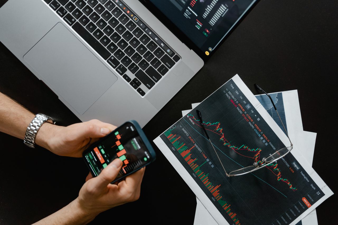Mains tenant un smartphone à côté d’un ordinateur portable affichant des graphiques boursiers, illustrant le trading numérique.